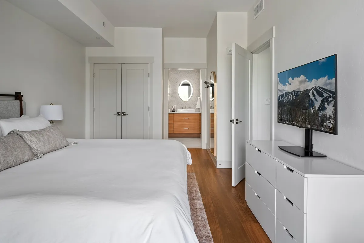 A serene master bedroom featuring a king-size bed with plush bedding, a modern dresser, a wall-mounted smart TV, and a glimpse of the bathroom entrance. The space is filled with natural light, creating a restful and inviting atmosphere.