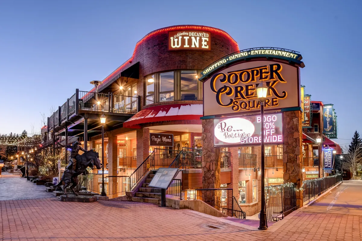 The Cooper Creek Square sign marks a popular downtown hub, with a wine shop entrance, outdoor seating, and festive decorative lighting creating a welcoming scene for dining, shopping, and entertainment.
