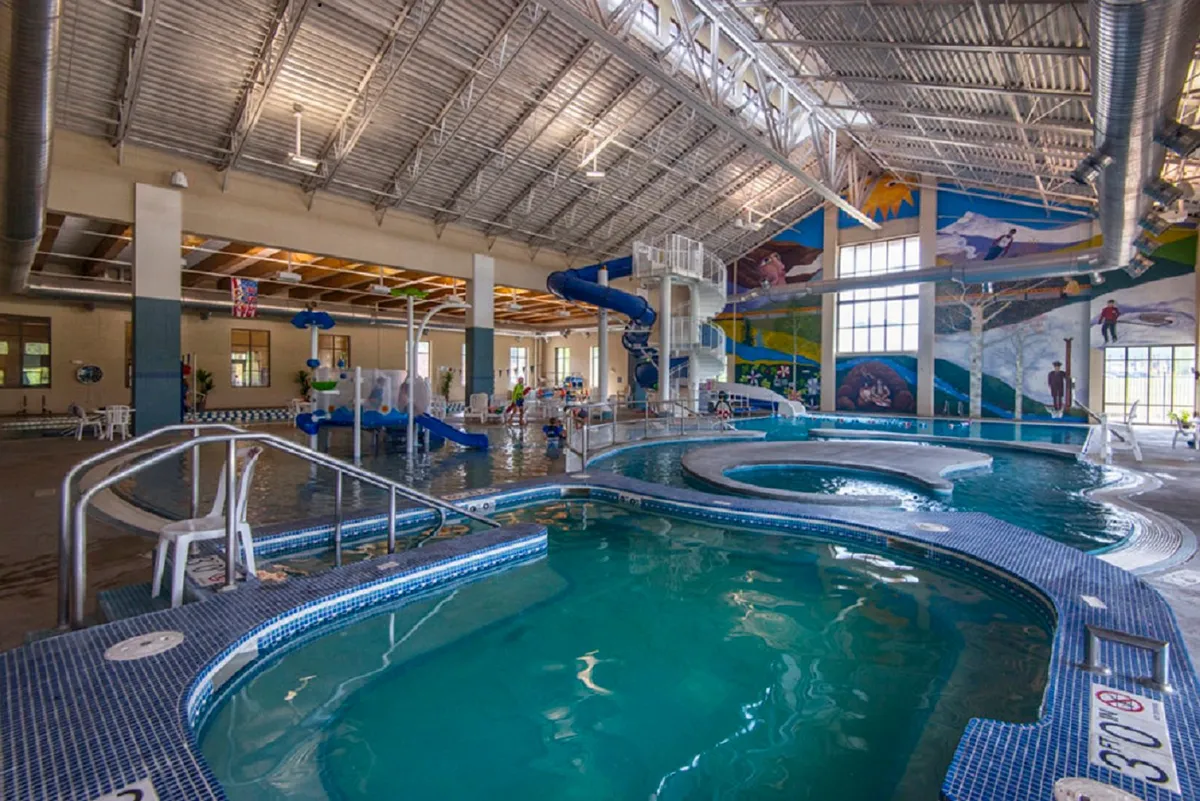 A lively indoor pool area featuring water slides, a swimming pool, murals, children playing, and lounge chairs. The atmosphere is energetic and joyful, capturing families and kids having fun.
