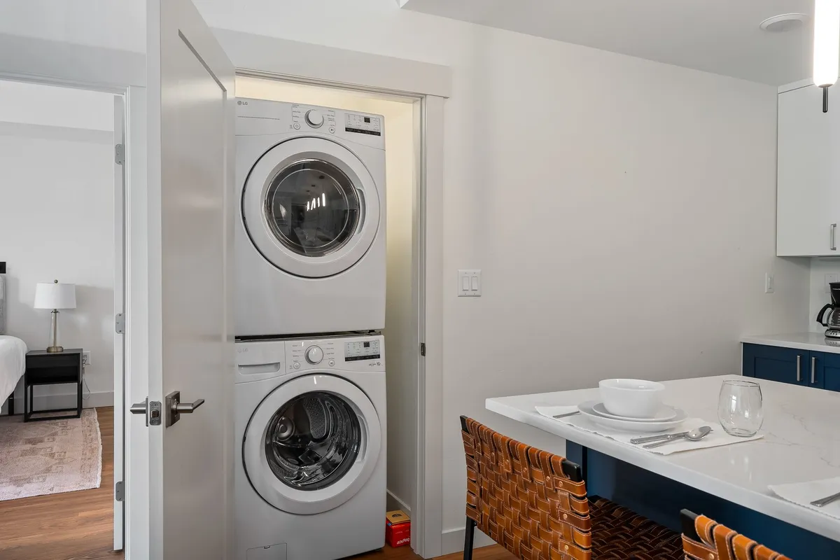 A contemporary laundry nook featuring a stacked washer and dryer next to a dining area with set tableware and a stylish, textured dining chair. The space is bright and efficient, emphasizing in-unit functionality for everyday needs.
