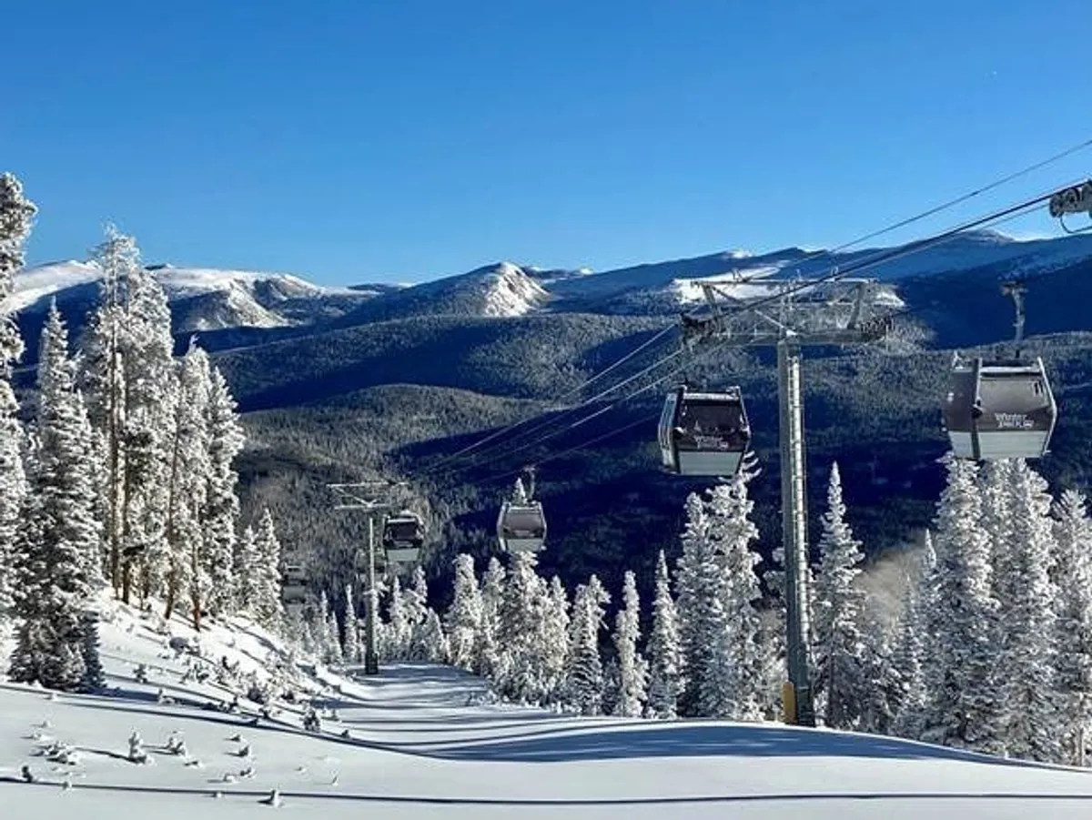 A sweeping exterior view featuring a gondola lift rising over snow-covered trees and a dramatic mountain range in the background. The scene is bustling with winter activity, hinting at skiing, snowboarding, and other alpine sports under clear blue skies.