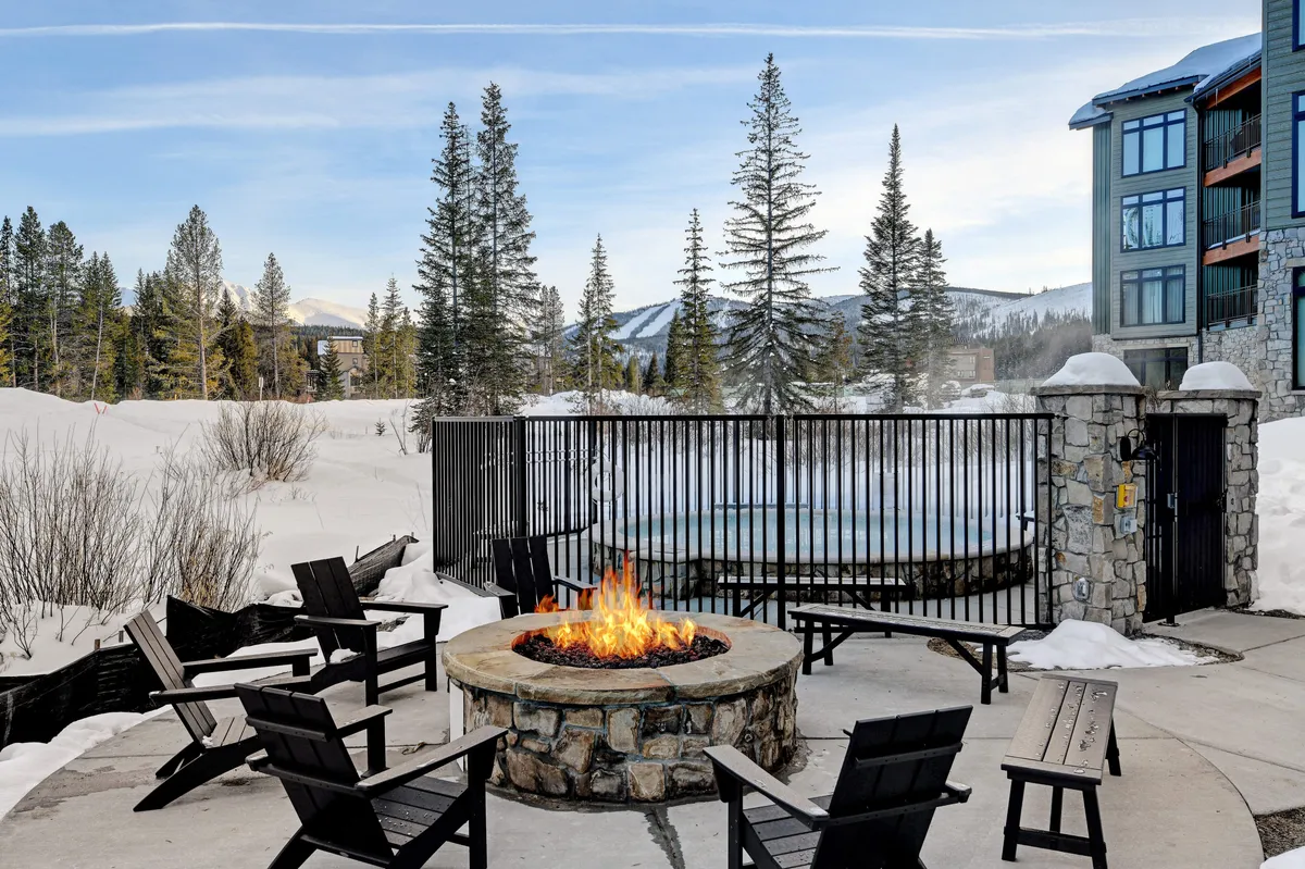 A cozy exterior setup with a fire pit surrounded by seating, a hot tub, snow-covered landscape, and mountain views. The scene suggests guests unwinding after a day of adventure, enjoying communal moments and the stunning Rocky Mountain setting.