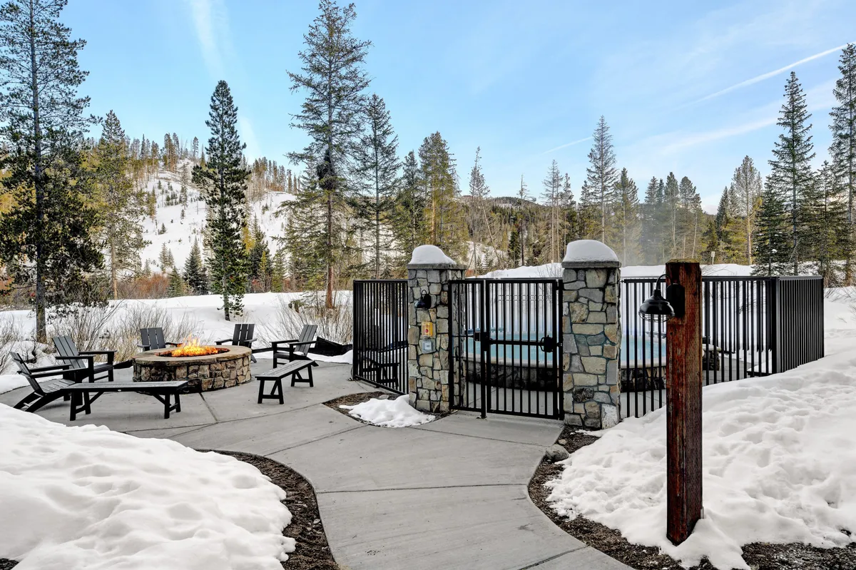 A cozy outdoor space featuring a fire pit and a hot tub enclosed by snow-laden pine trees. Guests gather around the fire, or relax in the steaming hot tub, all surrounded by a serene winter landscape that could transition to lush greenery in warmer months.