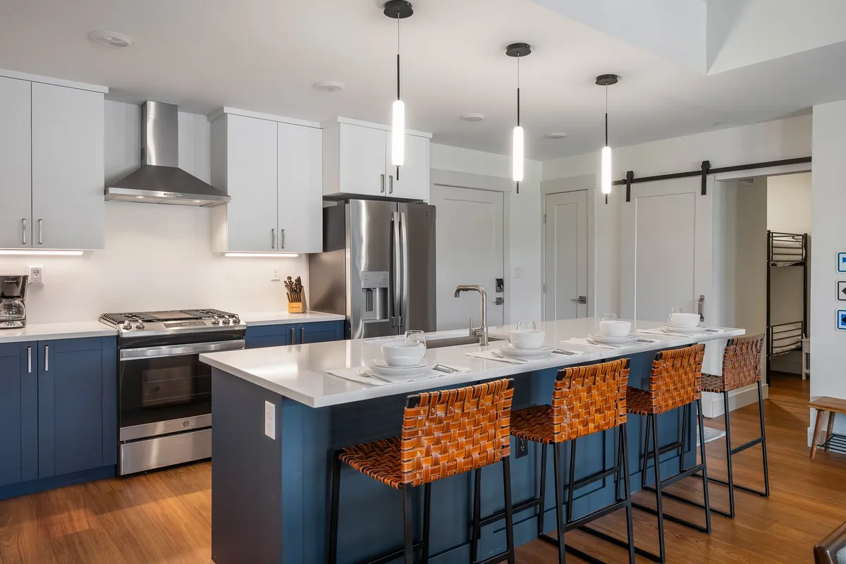 The image showcases a sleek, modern kitchen with a spacious island, inviting bar stools, and high-end stainless steel appliances—perfect for gathering, cooking, and socializing. The space is bright, contemporary, and designed to encourage both relaxation and conversation, hinting at memorable meals and gatherings.