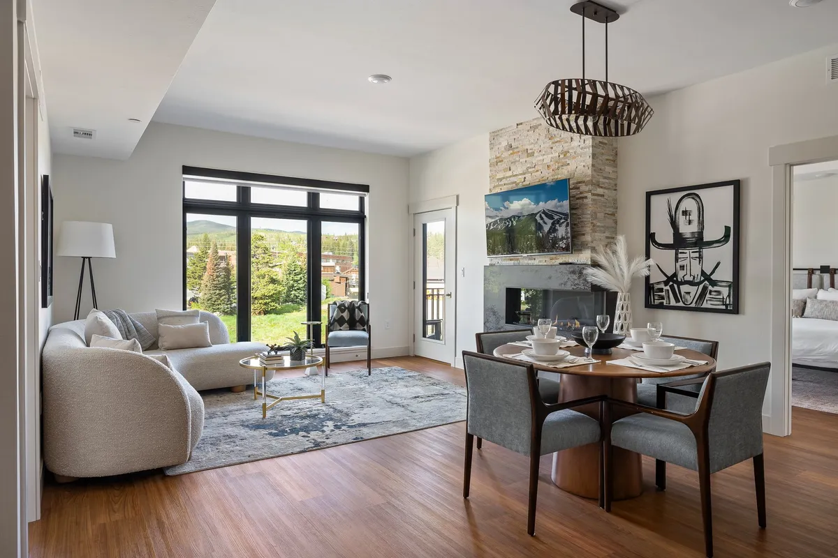 This image showcases an inviting, open-concept living area with a stylish sofa, a fireplace with a TV above it, a large dining table set for a meal, and expansive windows offering views of greenery outside. The overall ambiance is welcoming and comfortable, highlighting spaces for relaxation, entertainment, and social gatherings.