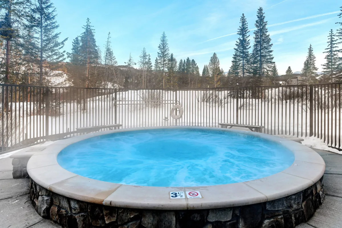 A steaming outdoor hot tub surrounded by snow and a rustic fence, with distant trees framing a serene winter landscape. The inviting scene suggests relaxation and socializing in all seasons, just steps from the residence.
