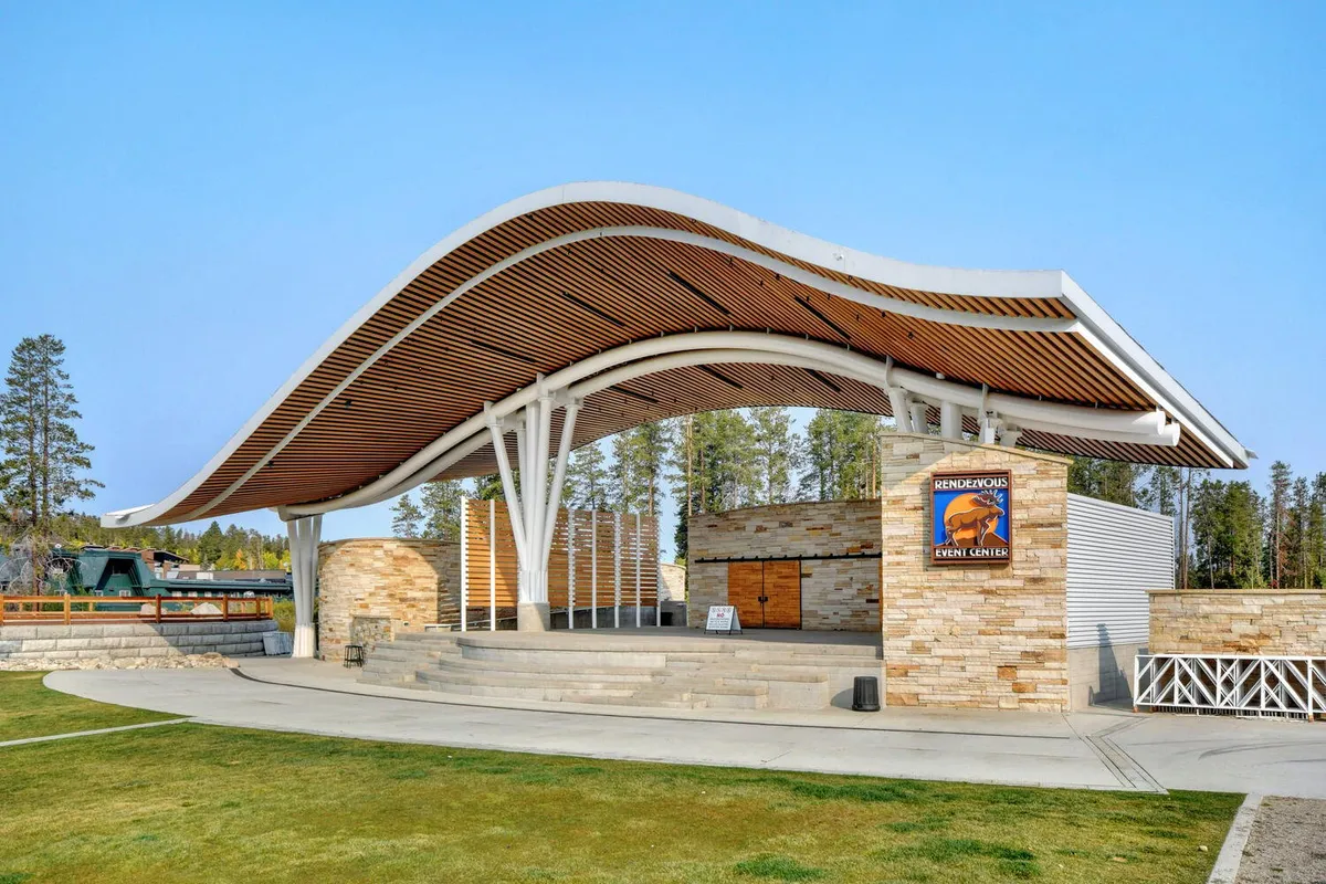 An outdoor amphitheater with a curved roof structure, stone walls, and steps leading to a stage, set in a scenic natural environment. The area is designed for community events, concerts, and theater performances.