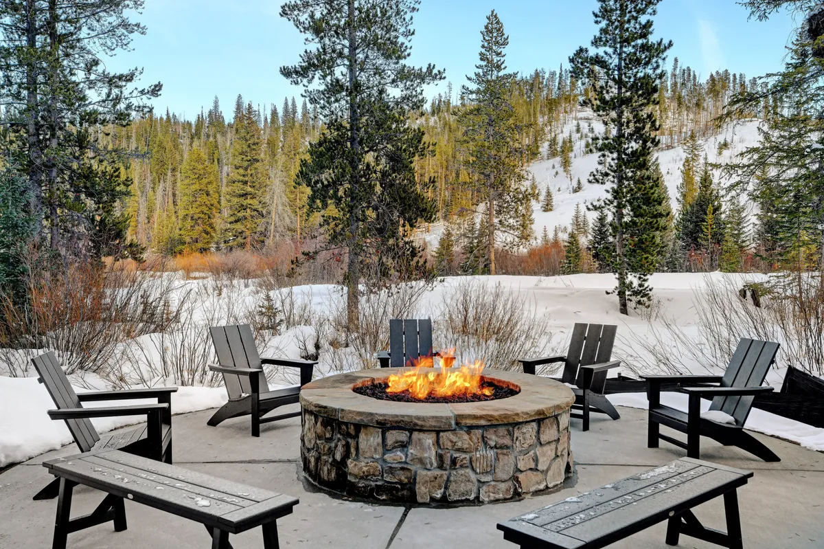 A cozy outdoor fire pit encircled by comfortable chairs sits amid a snowy landscape and towering pine trees. The fire glows warmly, inviting guests to gather, share stories, and enjoy the fresh mountain air.