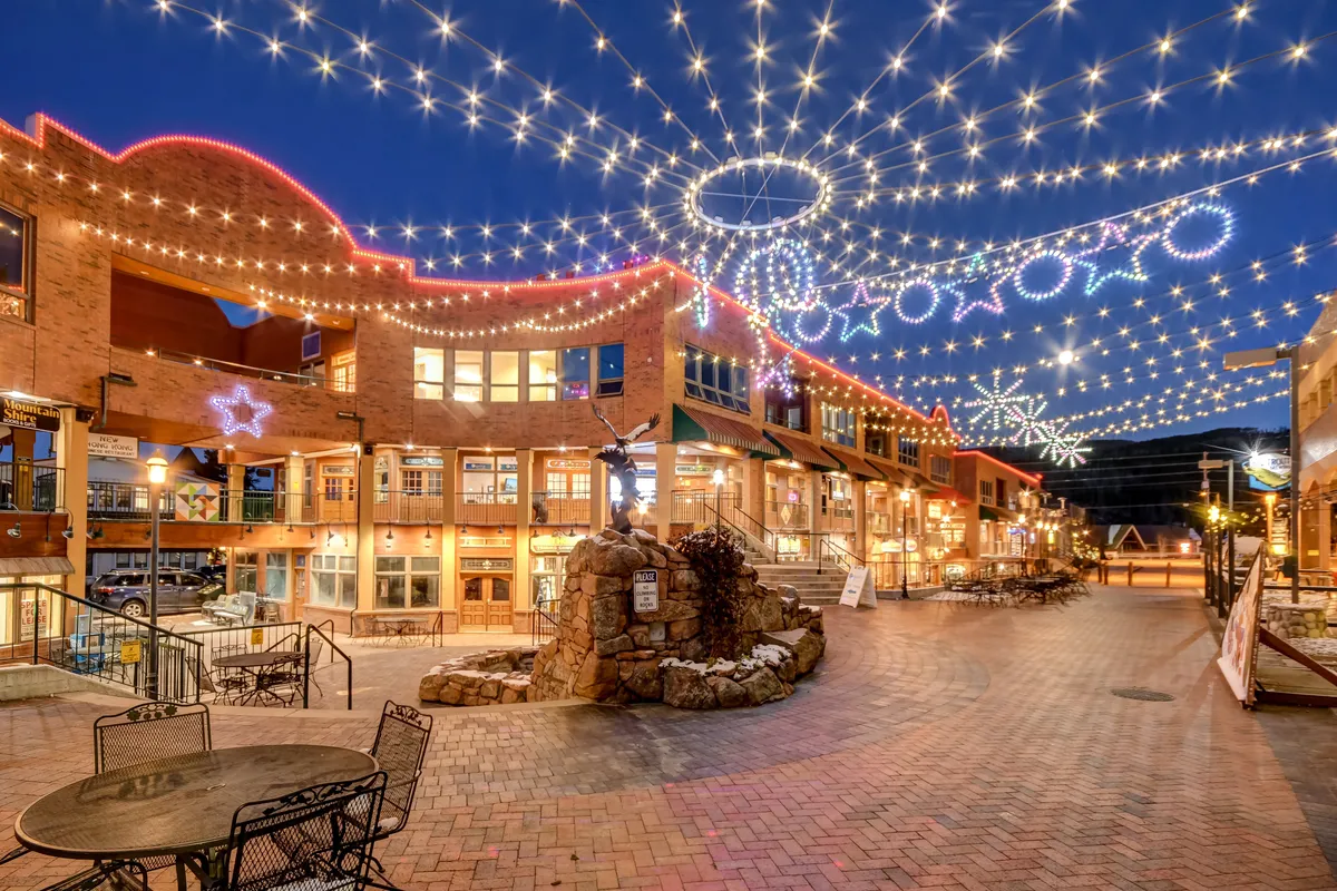 A vibrant, pedestrian-friendly downtown scene illuminated by overhead string lights, featuring decorative storefronts, a central fountain, and people enjoying shopping and dining. The ambiance is festive, lively, and inviting for all ages.